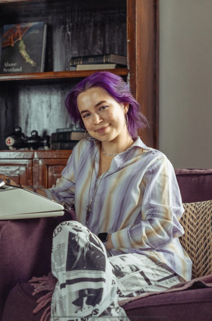 Heather Rose has purple hair and sits in an armchair with a typewriter